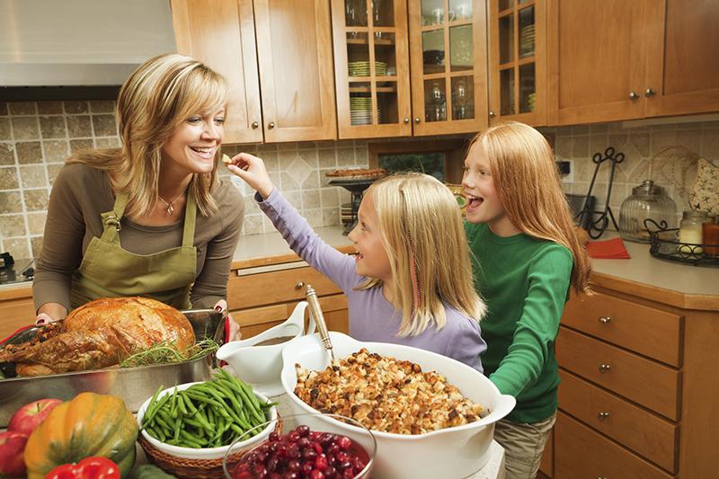 Two girls standing in a kitchen next to their mom. One of the girls is trying to feed her mom a piece of food. In front of the mom and girls is a table that has a turkey, bowl of green beans, casserole dish and bowl of cherries in it. - TAB Bank